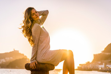 A young blonde in gray dress sitting in a fishing port on a beautiful sunset