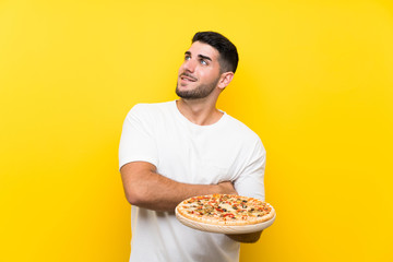 Young handsome man holding a pizza over isolated yellow wall looking up while smiling