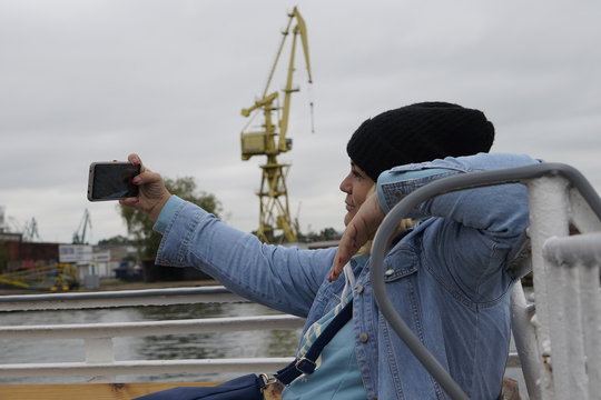 Gdansk, Poland - September 2019: A girl in a hat on the deck of a ship takes a selfie using a smartphone. A girl photographs herself against the background of ships in the port.