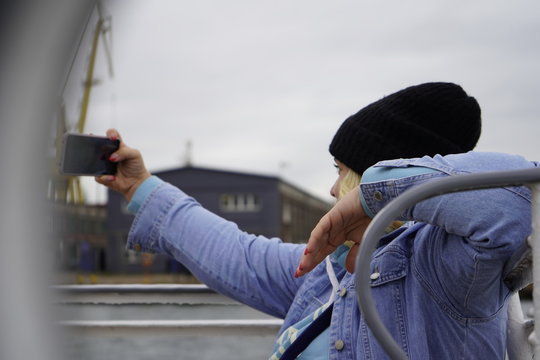 Gdansk, Poland - September 2019: A girl in a hat on the deck of a ship takes a selfie using a smartphone. A girl photographs herself against the background of ships in the port.