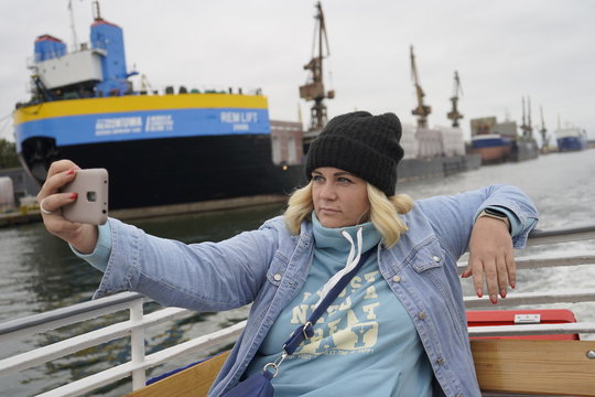 Gdansk, Poland - September 2019: A girl in a hat on the deck of a ship takes a selfie using a smartphone. A girl photographs herself against the background of ships in the port.