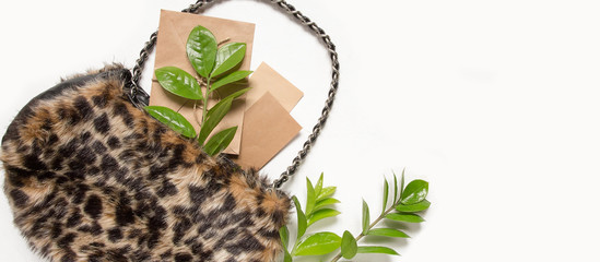 stylish bag leopard-patterned fur with green branches leaves, brushes, cosmetics, a glider, sunglasses light background. Top view. Fashionable bag female accessories. Summer fashion flat lay.