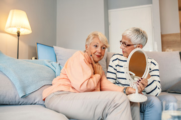 Senior women spending time together at home. Cheerful positive attractive senior lady friends looking at the mirror satisfied with nature beauty. Two cheerful senior women chatting at home.