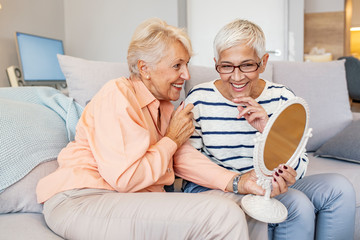 Senior woman examining her face in the mirror wit her friend. Two Senior Women Friends at home. Beautiful elderly woman holding mirror and talking with her friend.