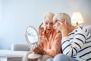 Senior woman examining her face in the mirror wit her friend. Two Senior Women Friends at home. Beautiful elderly woman holding mirror and talking with her friend.