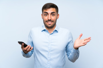 Young handsome man with his mobile over isolated blue wall with shocked facial expression