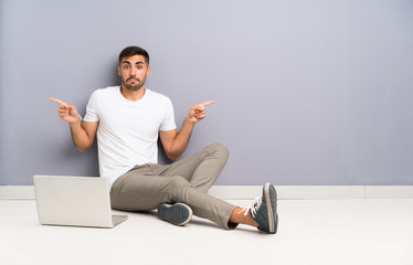 Young man with his laptop sitting one the floor pointing to the laterals having doubts