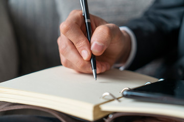 Close up of businessman hand taking notes in copybook with smartphone near it and blurred armchair in background. Concept of business lifestyle