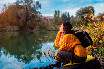 Traveler with backpack relaxing by autumn river at sunset. Young woman travels alone
