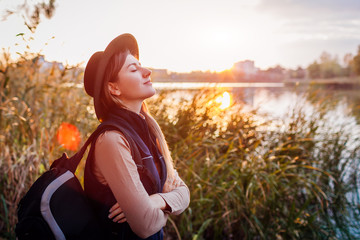 Traveler with backpack relaxing by autumn river at sunset. Young woman breathing deep feeling happy and free - obrazy, fototapety, plakaty