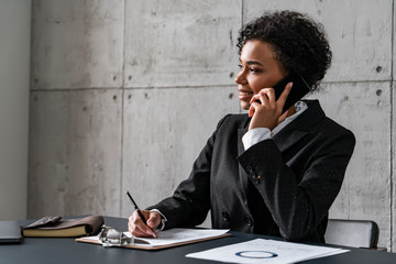 Portrait of beautiful smiling young African American businesswoman talking on smartphone and taking notes in blurred loft office. Concept of business lifestyle
