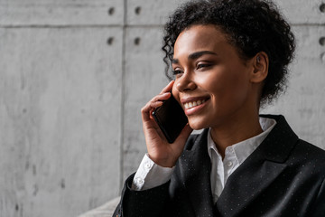 Attractive young smiling African American businesswoman in dark suit talking on smartphone in blurred loft office armchair. Concept of business communication