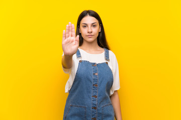 Young woman in dungarees over isolated yellow background making stop gesture