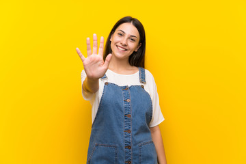 Young woman in dungarees over isolated yellow background counting five with fingers