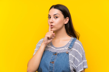 Young woman in dungarees over isolated yellow background doing silence gesture
