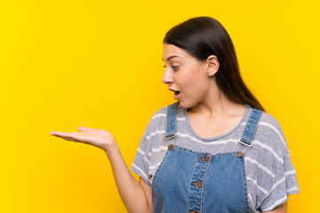 Young woman in dungarees over isolated yellow background holding copyspace imaginary on the palm
