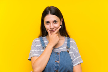 Young woman in dungarees over isolated yellow background thinking an idea