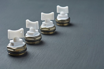 Row of four metal air valves with white plastic handle for to vent excess air and pressure from heater radiator in room lies on dark scratched desk in workshop. Space for text. Close-up