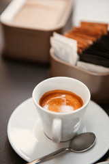 Close up. The small white ceramic mug (cup) with Italian espresso (coffee) on a table with a saucer and a metal spoon in a local Italian bar (coffee shop) in Milan, Lombardy, Italy. Beverage.