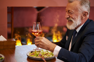 Senior elegant bearded and gray-haired man in tuxedo with glass of champagne sit in restaurant and laugh, look at person opposite. Background fireplace