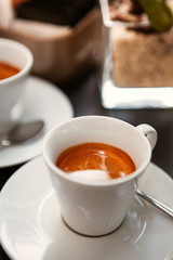 Close up. The small white ceramic mug (cup) with Italian espresso (coffee) on a table with a saucer and a metal spoon in a local Italian bar (coffee shop) in Milan, Lombardy, Italy. Beverage.