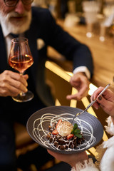 Close image of hand with glass of champagne, sweet dessert on grey dish. Beared man in tuxedo drinking