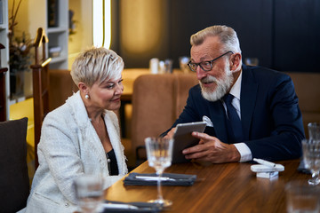 Beautiful senior couple in love speaking in rich restaurant, man show smith interesting in laptop. Modern couple