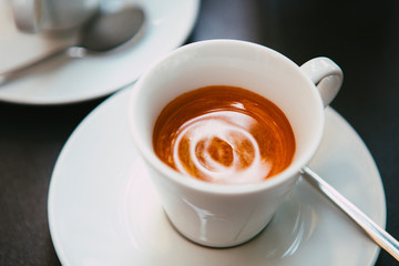 Close up. The small white ceramic mug (cup) with Italian espresso (coffee) on a table with a saucer and a metal spoon in a local Italian bar (coffee shop) in Milan, Lombardy, Italy. Beverage.
