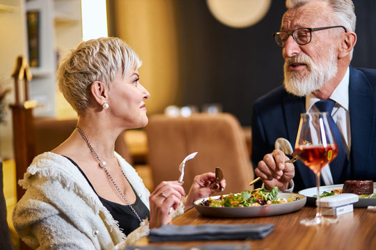 Elderly elegant people taking meal in beautiful restaurant. Free and modern, using iqos, e-cigarettes. Caucasian grey-haired and beared male in tuxedo and glasses. Woman looks at him and listen