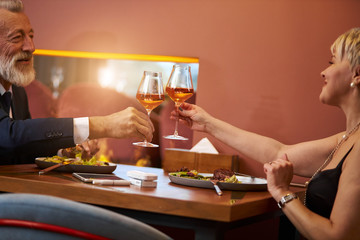 Grey-haired man in tuxedo and senior lady in black dress clink glasses with champagne in expensive restaurant. Look and happily smile. Romantic atmosphere