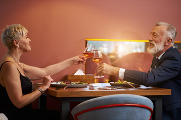 Senior elegant couple sit in restaurant and clink glasses with champagne, celebrate. Bearded gray-haired man in tuxedo and caucasian woman in black dress. Love, relationship, restaurant concept