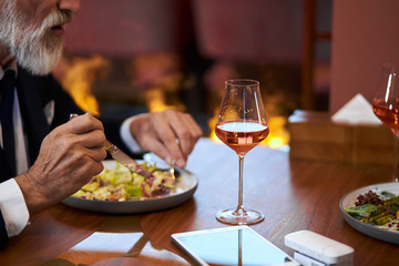 Bearded gray-haired man in elegant restaurant.Close photo of mature hands. Glass of champagne and dish of salad on table.