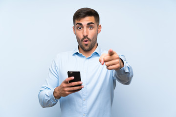 Young handsome man with his mobile over isolated blue wall surprised and pointing front