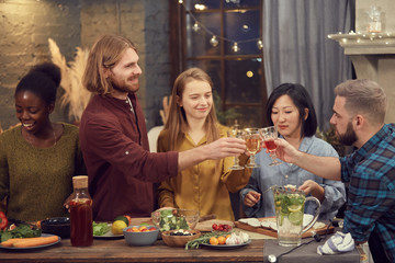 Multi-ethnic group of young people enjoying party together standing at table in modern interior and clinking glasses