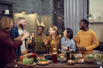 Multi-ethnic group of smiling young people enjoying dinner together standing at table in modern...