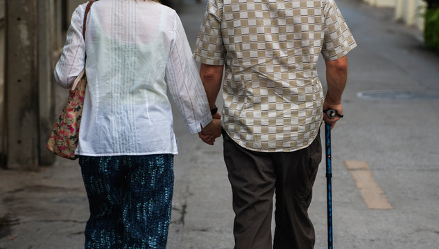 Back View Of Well-dressed Senior Couple Aged Man And Woman In Hats Walking Arm-in-arm In Town.