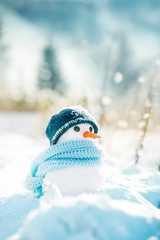 Little snowman made of snow in a knitted hat and a blue scarf on a background of a winter snowy...