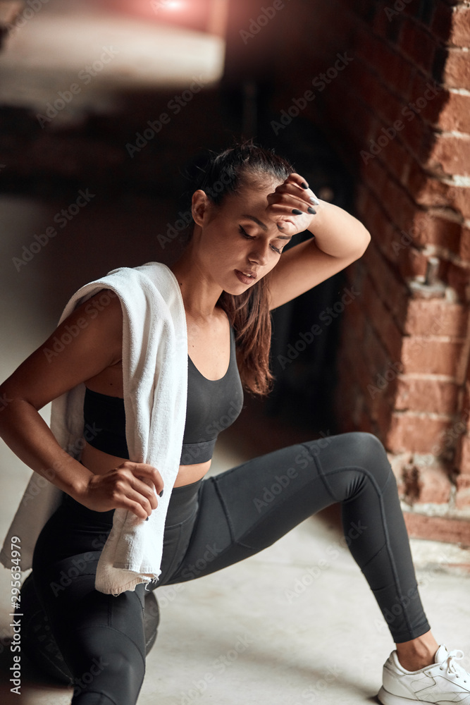 Wall mural Fitness girl tired and sit on floor in gym. Dressed in black sportswear, white towel on shoulders. Brickwall background