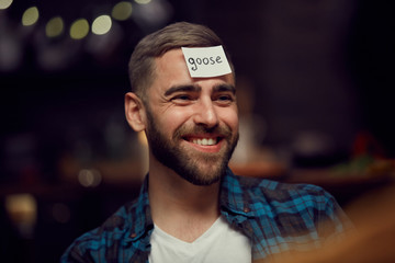 Head and shoulders portrait of smiling bearded man with goose label on forehead playing guessing game with friends during party