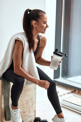 Positive, smiling fitness female in sportswear sit with plastic bottle after fitness. Have rest,...