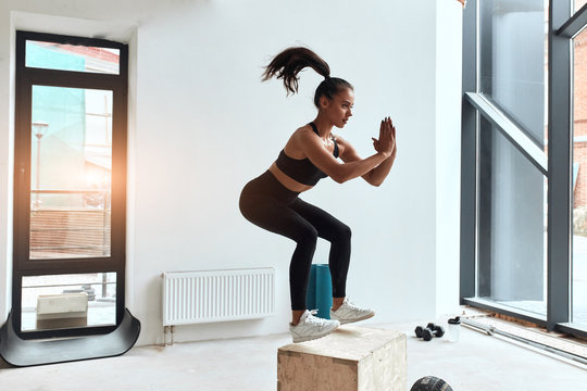 Box jumping in process at crossfit style in gym, box jump performing of young fit girl. Window background