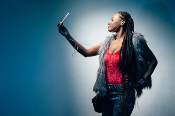 Dark-skinned model listens to music and takes a selfie. Girl with braids taking pictures of himself on the phone. African-American woman in a fur vest. African woman with a smartphone.