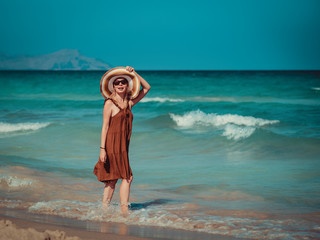 Young blond European woman in colorful sun hat standing against wonderful blue sea.  She enjoying her rest at seacoast and looking towards sea.
