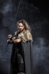 Man dressed in medieval armor and raincoat with longs word over smoke background. Courage fantasy warrior knight with long hair concept historical photo