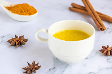 Golden (turmeric) milk in a white cup on the marble background. A spoon with turmeric powder.  Cinnamon and anise. 