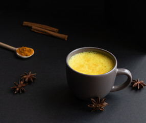 Golden (turmeric) milk in a gray cup on the dark background. A spoon with turmeric powder. Cinnamon and anise. 