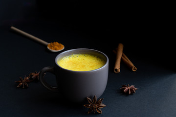 Golden (turmeric) milk in a gray cup on the dark background. A spoon with turmeric powder. Cinnamon and anise. 
