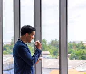 A young Asian businessman drinks coffee in the office window in a comfortable