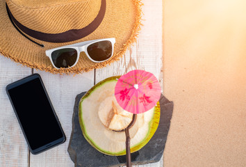 Fresh coconut drink and summer accessories, sunglasses, hat, mobile phone on white wood table at sand beach with sunlight