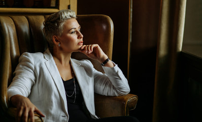 Pleasant looking delighted female in white glorious jacket, with thoughtful expression leans on armrest of fashion armchair, keeps chin, doesnt look to camera